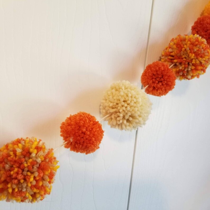 May include: A garland of orange, yellow, and cream yarn pom-poms strung together with white thread.