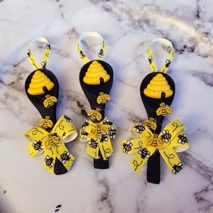 May include: Three wooden spoons painted black with a yellow beehive and bees design. Each spoon has a yellow bow with a bee pattern. The spoons are hanging from a white string.