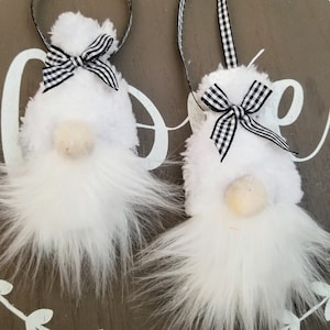 May include: Two white plush gnome ornaments with fluffy beards and small beige noses. Each gnome has a black and white checkered bow and a black ribbon for hanging. The ornaments are set against a gray background.
