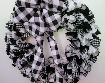 May include: A circular wreath constructed from numerous fabric strips in black, white, and black-and-white buffalo check patterns. A large, prominent bow made from the same black-and-white buffalo check ribbon is attached to the top center.