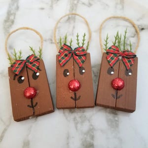 May include: Three wooden reindeer ornaments with brown bodies, red glitter noses, and green and red plaid bows. Each ornament has a jute handle and faux greenery. The reindeer have painted eyes and smiles.