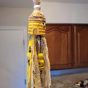 May include: A decorative hanging ornament featuring a yellow beehive design with a twine-wrapped top. The ornament includes wooden beads and a fabric tassel with bee and honeycomb patterns. The overall design is approximately 10 inches in length.