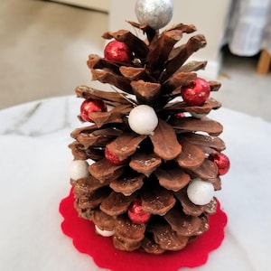 May include: A handcrafted Christmas tree ornament made from a pine cone. The brown pine cone is decorated with small red and white ball ornaments and a silver glitter ball at the top. The ornament sits on a red felt base.