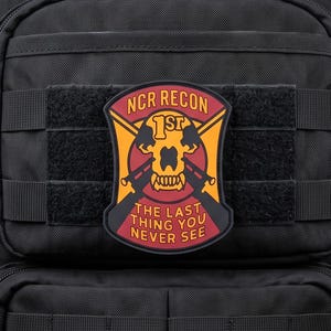 Könnte beinhalten: Ein schwarzer taktischer Rucksack mit einem Aufnäher auf der Vorderseite. Der schildförmige Aufnäher hat einen gelben Hintergrund, einen roten Rand und ein Totenkopfmotiv. Der Text lautet NCR RECON 1ST THE LAST THING YOU NEVER SEE.