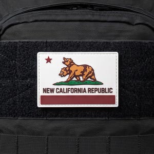 Könnte beinhalten: Ein schwarzer taktischer Rucksack mit einem rechteckigen Aufnäher der Flagge der New California Republic. Der Aufnäher zeigt einen braunen Bären, einen roten Stern und den Text NEW CALIFORNIA REPUBLIC über einem roten Streifen auf weißem Grund.