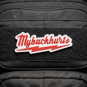 May include: A black tactical backpack featuring a rectangular patch on the upper front pocket. The patch displays the white and red text Mybackhurts in a stylised font with a lightning bolt graphic underneath, set against a black textured background.