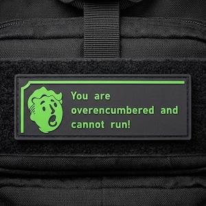 May include: A rectangular black patch with a green border and green text that reads "You are overencumbered and cannot run!" next to a green graphic of a surprised cartoon face. The patch is attached to the front of a black tactical backpack.