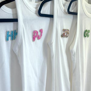 May include: White ribbed tank tops hanging on black hangers. Each tank top has a different set of colorful beaded letters: HH, PJ, a multi-colored design, and GC. The tank tops are made of a soft, ribbed material.