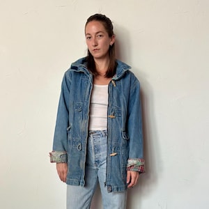 Może przedstawiać: Jasnoniebieska kurtka dżinsowa z kapturem i drewnianymi zapięciami na kołki. Kurtka ma kraciastą podszewkę na mankietach. Osoba ma na sobie jasnoniebieskie dżinsy i czerwone tkane buty.