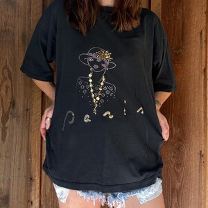 May include: Black t-shirt featuring a rhinestone design of a woman in a hat and the word "Paris" in gold lettering. The shirt has short sleeves and a relaxed fit.