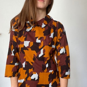 Könnte beinhalten: Ein langärmeliges Kleid mit Kragen. Das Kleid ist aus einem Stoff mit braunem, orangefarbenem und weißem geometrischem Muster.