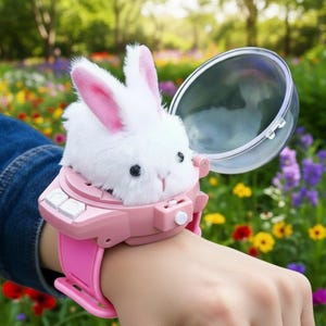 Puede incluir: Un reloj de juguete rosa y blanco con un conejito blanco y esponjoso con orejas rosas. El reloj tiene una cúpula transparente y una correa rosa. El fondo es un jardín borroso con flores coloridas.