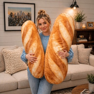 May include: Two large, plush baguette pillows with realistic crust details are held by a person. The pillows are primarily tan and brown, resembling freshly baked bread. The person is wearing a blue sweater and jeans.