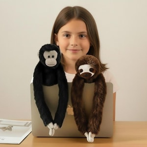 Pode incluir: Um macaco preto e uma preguiça castanha de pelúcia com braços longos, pendurados em um laptop. O macaco tem rosto e mãos brancas, enquanto a preguiça tem rosto e pés brancos. Os brinquedos estão na frente de uma menina.
