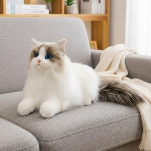 May include: A soft toy cat with white fur, blue eyes, and brown markings sits on a grey sofa. The cat has a long, fluffy tail with grey and brown fur. A cream-coloured blanket is draped over the sofa.