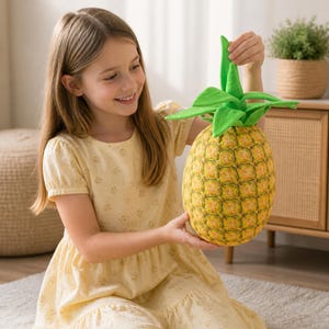May include: A plush pineapple toy with a textured yellow and green exterior. The toy has a green leafy top and is being held by a person wearing a yellow dress. The pineapple is a fun, decorative item.