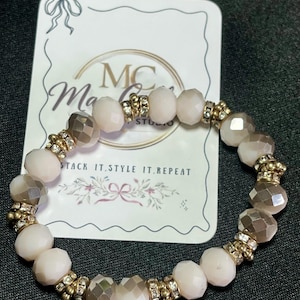 May include: A beaded bracelet featuring alternating faceted and round beads in shades of pink and gray. Gold-toned spacers with clear rhinestones separate the beads. The bracelet is displayed on a card that reads "Stack it. Style it. Repeat."
