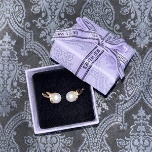 Peut inclure: Une paire de boucles d'oreilles dorées avec une perle blanche et une petite pierre transparente, présentées dans une boîte carrée doublée de velours noir. La boîte est violet clair avec un ruban assorti portant l'inscription BEST WISHES.