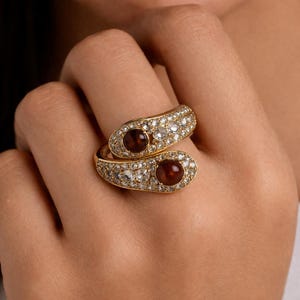 Pode incluir: Um anel dourado com um design envolvente. O anel apresenta duas secções ovais, cada uma com uma pedra grande, redonda, castanho-avermelhada. As secções são incrustadas com pequenas pedras preciosas transparentes, criando um efeito brilhante.