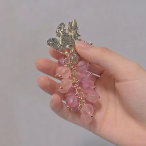 Puede incluir: Una pinza para el pelo con forma de uva con detalles en rosa y dorado. La pinza presenta un racimo de cuentas rosas, que se asemejan a uvas, suspendidas de un tallo de metal dorado con detalles de hojas. La pinza se sostiene en una mano.