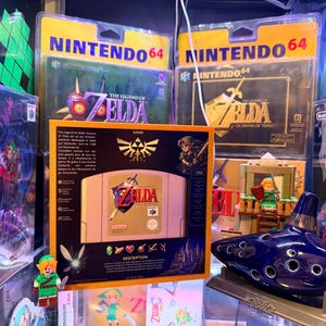 Op de afbeelding: Een verzameling The Legend of Zelda Nintendo 64 videospellen, inclusief een verpakte Ocarina of Time cartridge met een goudkleurige lijst, een blauwe keramische ocarina en een klein Link-figuurtje, allemaal uitgestald tussen diverse speldozen.