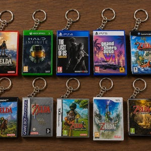May include: A collection of ten miniature video game case keychains featuring various titles including The Legend of Zelda, Halo Infinite, The Last of Us, and Grand Theft Auto VI. Each rectangular keychain includes a metal ring and chain attachment.