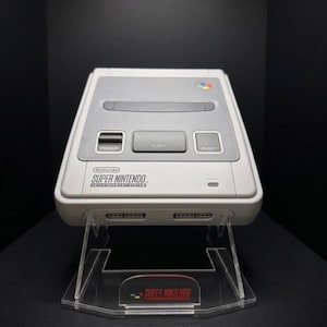 Peut inclure: Une console Super Nintendo Entertainment System gris clair, présentée sur un présentoir en acrylique transparent. La console affiche le logo Super Nintendo et des boutons d'alimentation, d'éjection et de réinitialisation. Le présentoir porte également le logo Super Nintendo.