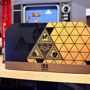 Könnte beinhalten: Eine Nintendo Switch Konsolen-Dockingstation mit einem schwarz-goldenen geometrischen Muster. Die Vorderseite zeigt ein großes zentrales Dreieck mit dem Triforce-Symbol und kleineren kreisförmigen Icons sowie dem Nintendo Switch Logo unten mittig.