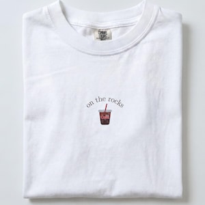 Op de afbeelding: Een wit T-shirt met korte mouwen en een ronde hals. In het midden op de borst staat een kleine geborduurde afbeelding van een donker drankje in een plastic beker met een rood rietje, vergezeld door de zwarte tekst "on the rocks".