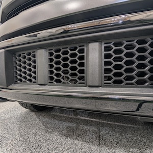 May include: Close-up of a black truck bumper with a chrome trim. The bumper has a honeycomb grille in the middle and rectangular grilles on the sides. The front of the vehicle is visible.