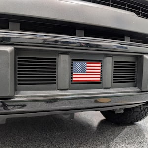 May include: A black vehicle front bumper featuring a horizontal grille with three distinct sections. The centre section of the grille displays a rectangular emblem of the United States flag with its characteristic red and white stripes and blue field.