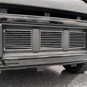 May include: Close-up of a vehicle's front bumper. The bumper is grey with a chrome accent and features three rectangular air vents with horizontal slats. A portion of a tyre is visible.