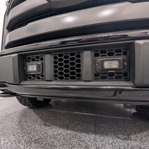 May include: A black vehicle front bumper featuring a central honeycomb grille insert with two rectangular LED auxiliary lights mounted symmetrically on either side. The assembly is finished in a matte and glossy black texture with visible hardware.