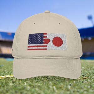 Op de afbeelding: Beige baseballpet met een geborduurd ontwerp met de Amerikaanse en Japanse vlaggen. De Amerikaanse vlagzijde heeft een rood hart en de Japanse vlagzijde heeft een rode cirkel. De pet staat op een groen grasveld.