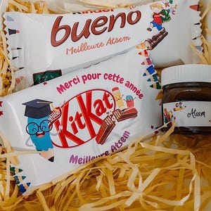 Peut inclure: Un coffret cadeau contenant une barre chocolatée Kinder Bueno, une barre Kit Kat et un pot de pâte à tartiner au chocolat. L'emballage présente des illustrations colorées sur le thème de l'école et du texte en français. La boîte est remplie de paille décorative.