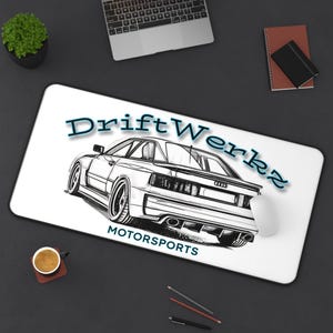 May include: A rectangular desk mat with a white surface featuring a black and white line art illustration of a sports car. The text DriftWerkz is printed in blue above the car, and the word MOTORSPORTS is printed in black below the car.