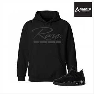 Puede incluir: Sudadera con capucha negra con la palabra "Rare" en fuente cursiva y "AIR" debajo. Una zapatilla negra está en primer plano. La sudadera con capucha tiene un bolsillo delantero y una capucha. La zapatilla es negra con suela negra.