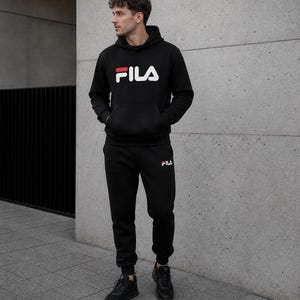 Puede incluir: Conjunto de ch&aacute;ndal negro FILA, compuesto por una sudadera con capucha y un pantal&oacute;n de ch&aacute;ndal. El logotipo de FILA blanco y rojo es visible en ambas prendas. La persona lleva zapatillas negras. Adecuado para ropa informal.