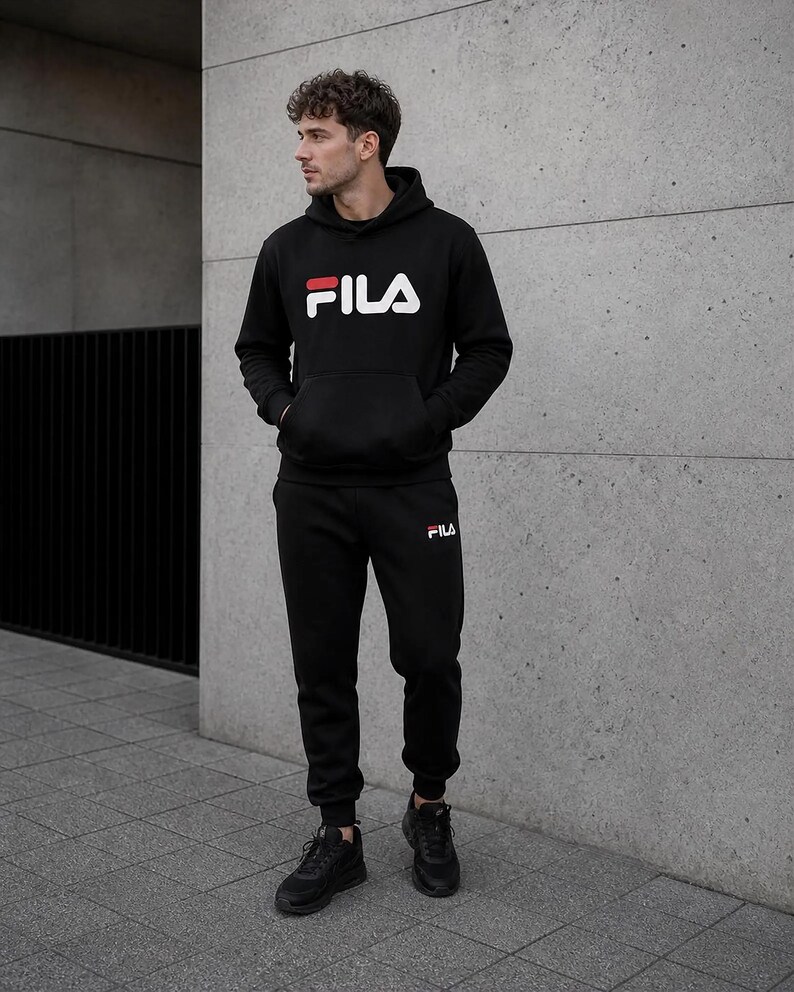 Puede incluir: Conjunto de ch&aacute;ndal negro FILA, compuesto por una sudadera con capucha y un pantal&oacute;n de ch&aacute;ndal. El logotipo de FILA blanco y rojo es visible en ambas prendas. La persona lleva zapatillas negras. Adecuado para ropa informal.