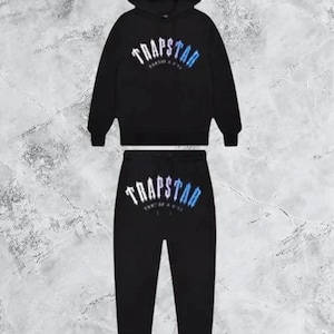 May include: Black tracksuit set featuring a hooded sweatshirt and matching sweatpants. The word "TRAPSTAR" is printed across the chest and on the pants in white and blue. The set is displayed on a marble-like background.