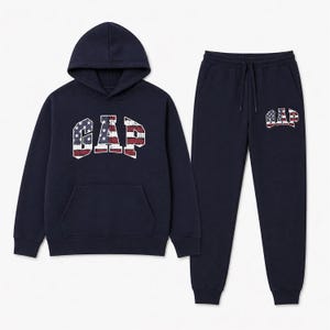 Könnte beinhalten: Marineblaues Hoodie- und Jogginghosen-Set. Der Hoodie hat eine Vordertasche und eine Kapuze. Das Wort "GAP" ist im Used-Look mit einem rot-weiß-blauen Flaggenmuster auf der Vorderseite aufgedruckt. Die Jogginghose hat ein passendes Logo.