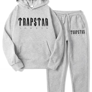May include: A light gray hoodie and sweatpants set. The hoodie has a front pocket and a hood. The word "TRAPSTAR LONDON" is printed in black on the front of the hoodie and on the left pant leg. The drawstring pants have pockets.