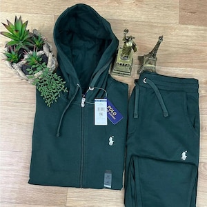 Op de afbeelding: Een donkergroene trainingspakset, bestaande uit een hoodie met ritssluiting en een bijpassende joggingbroek. De hoodie heeft een capuchon met trekkoord en een klein logo op de borst. De joggingbroek heeft een tailleband met trekkoord en een bijpassend logo.