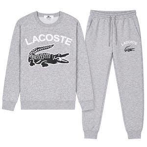 Op de afbeelding: Een grijze Lacoste sweatshirt en bijpassende joggingbroek set. De sweatshirt heeft het Lacoste logo en een zwarte krokodil afbeelding. De joggingbroek heeft ook het Lacoste logo. De set is gemaakt van een zacht, comfortabel materiaal.