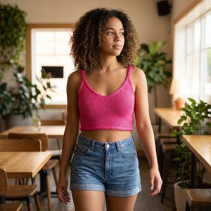May include: A woman wearing a bright pink knitted crop top and denim shorts. The top has a V-neck and spaghetti straps. The shorts are a light blue wash. The woman is standing in a cafe with wooden tables and chairs.