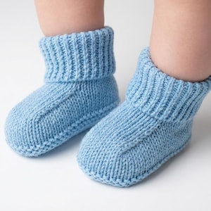 May include: A pair of light blue knitted baby booties. The booties have a ribbed cuff and a rounded toe. The soft, cozy knitwear is designed to keep a baby's feet warm. The booties are shown on a white background.