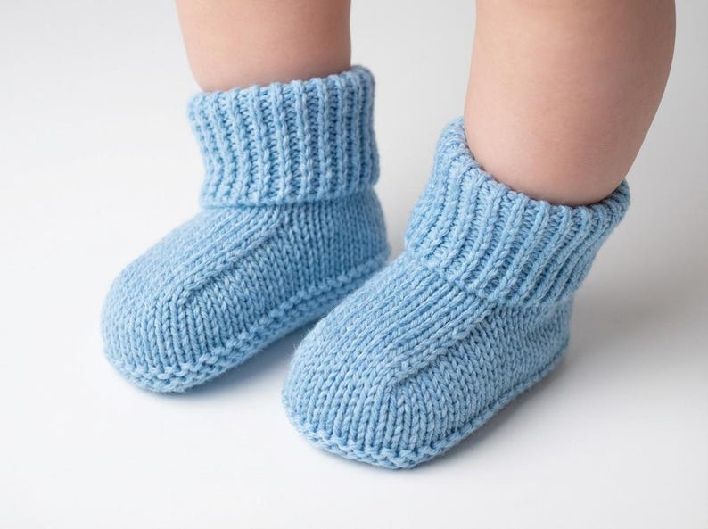 May include: A pair of light blue knitted baby booties. The booties have a ribbed cuff and a rounded toe. The soft, cozy knitwear is designed to keep a baby's feet warm. The booties are shown on a white background.