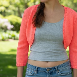 Könnte beinhalten: Ein korallenfarbener Bolero-Pullover mit kurzen Ärmeln. Das gecroppte Strickteil hat einen runden Ausschnitt und wird über einem grauen Tanktop und einer Jeans getragen. Der Hintergrund ist eine verschwommene Szene im Freien mit grünem Gras und Bäumen.