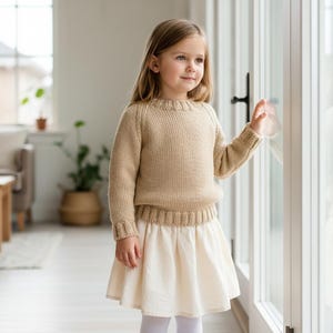 May include: A young girl wearing a beige knit sweater and a cream-colored skirt stands near a glass door. The sweater has a ribbed neckline and cuffs. The skirt is flared. The background shows a living room with a sofa and a wooden table.