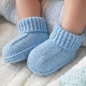 May include: A pair of light blue knitted baby booties. The booties have a ribbed cuff and a simple, classic design. They are shown on a soft, textured blanket, suggesting comfort and warmth.